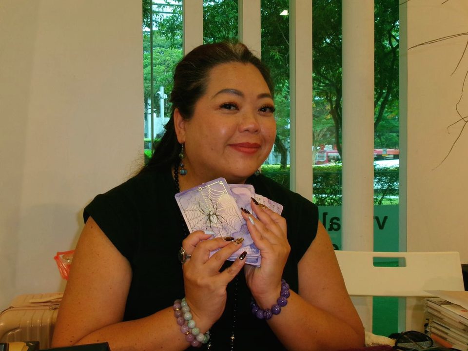 Perilla at her table at a fair, holding up a few of her cards